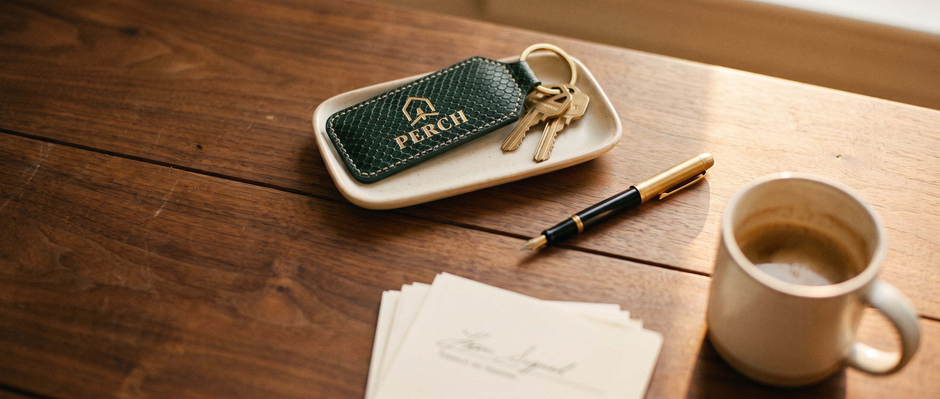 PERCH Agents — activate your PERCH partner account. Brass keys with a PERCH leather key fob rest on a warm walnut closing table at golden hour, alongside signed papers, a fountain pen, and morning coffee.
