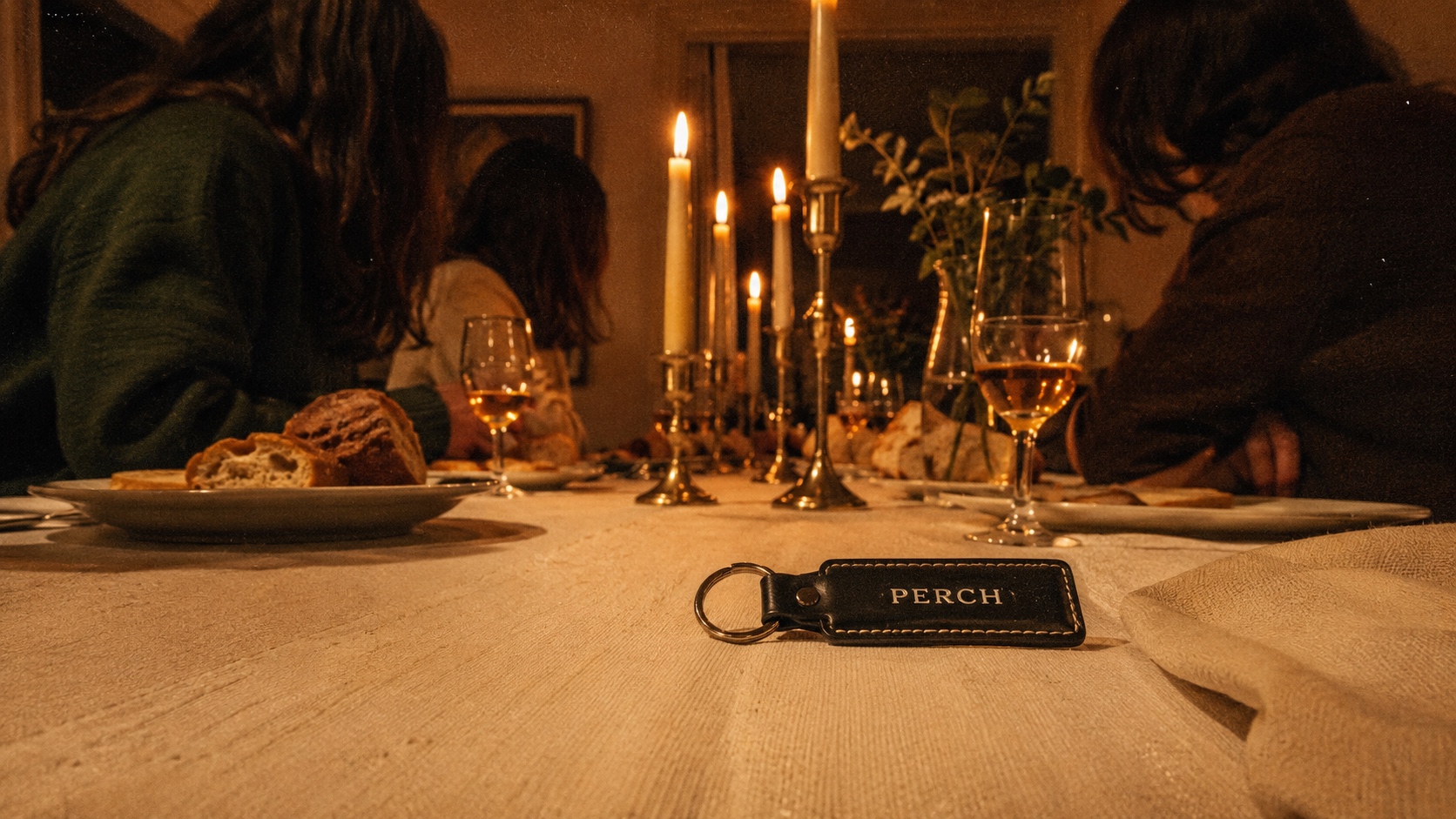A long candlelit dinner table, adults in profile with faces obscured, a small PERCH keyfob on the corner
