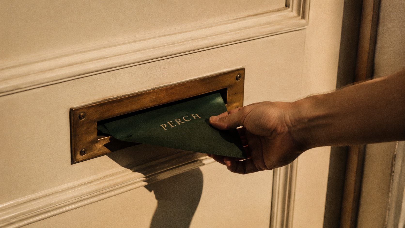 A hand slipping a small envelope with a forest wax seal into a brass mail slot on a warm cream-painted door